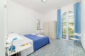 A hotel room with a bed, a desk with a chair, a wardrobe, and a view of the outdoors through a glass door. - Residence Le Villette (Ravello)