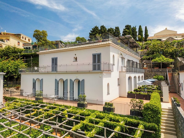 A two-story building with a balcony, arched windows, and a garden with a trellis.