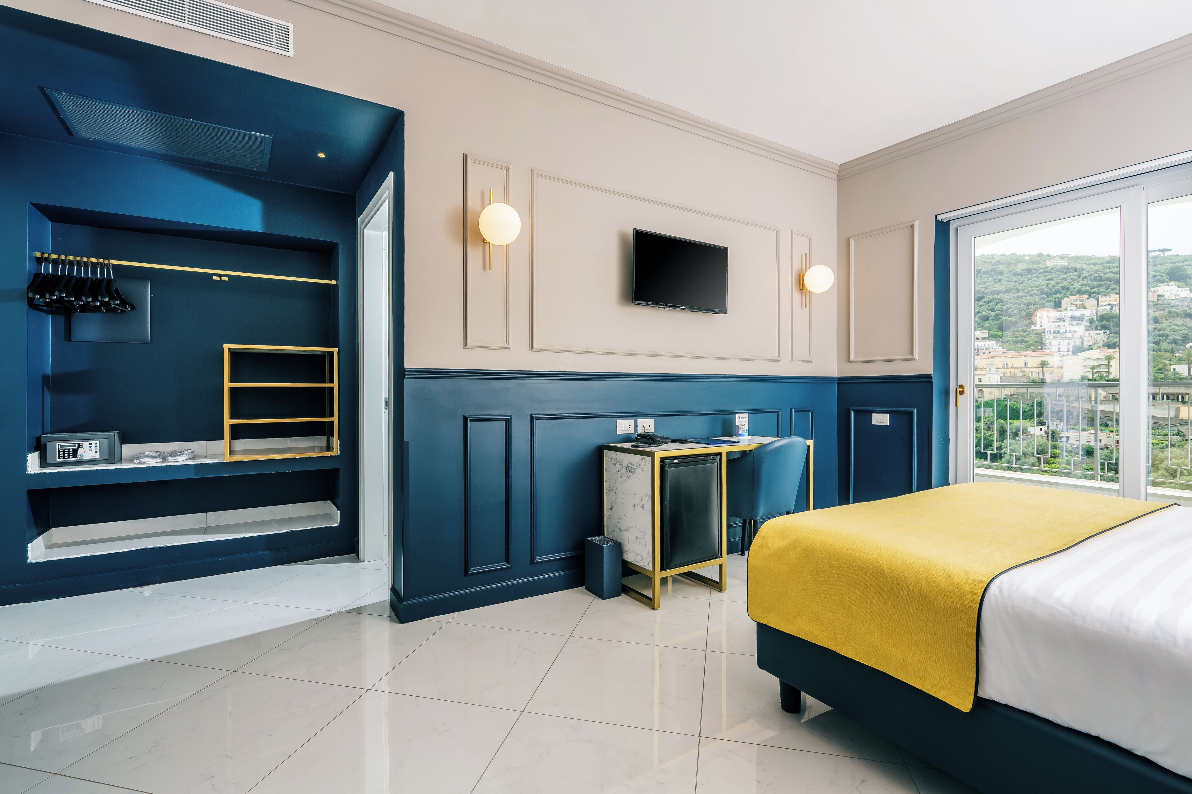 A modern bedroom with a bed, a desk, a TV, and a balcony with a city view.