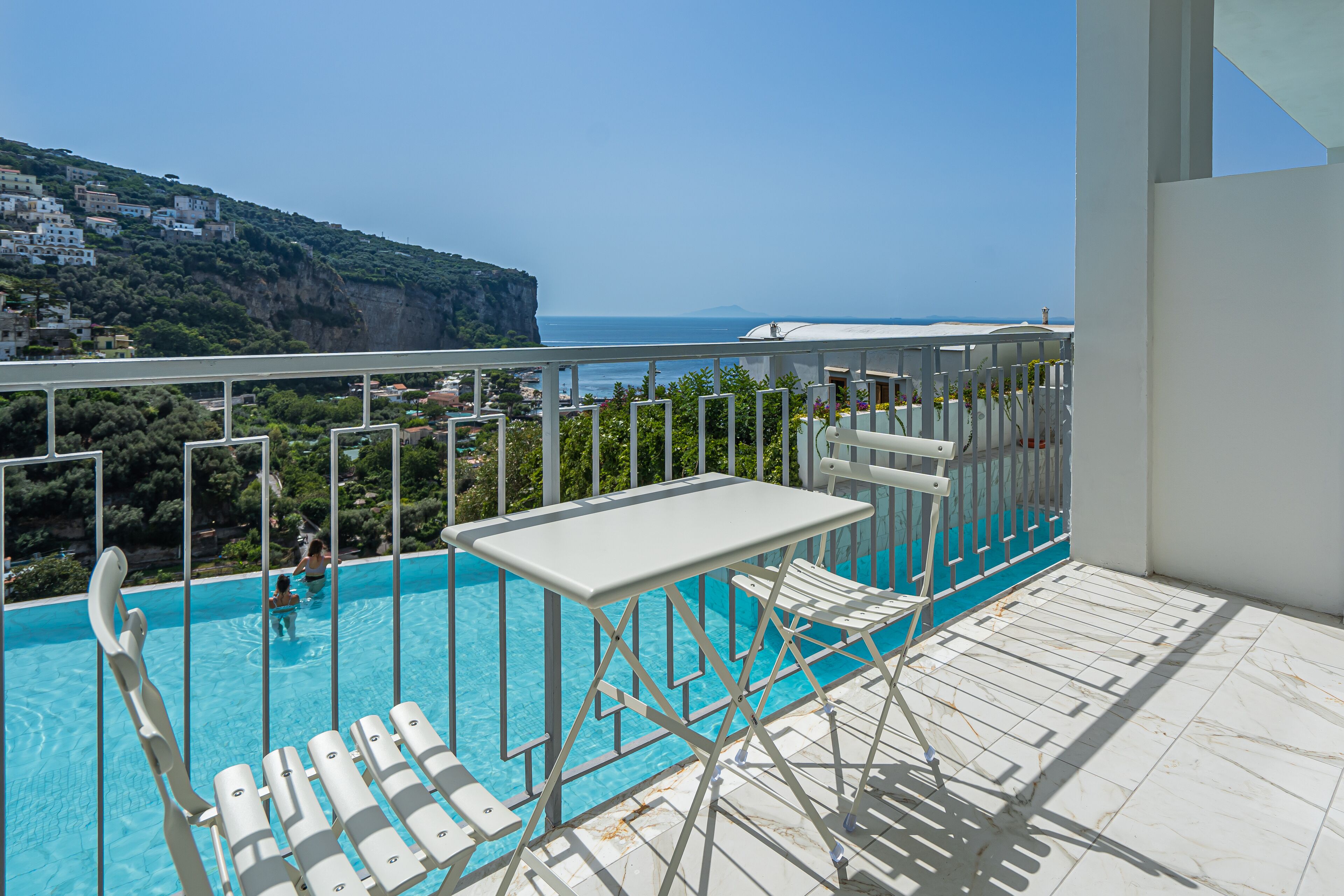 A balcony with a pool, white furniture, and a view of the sea and a hillside.