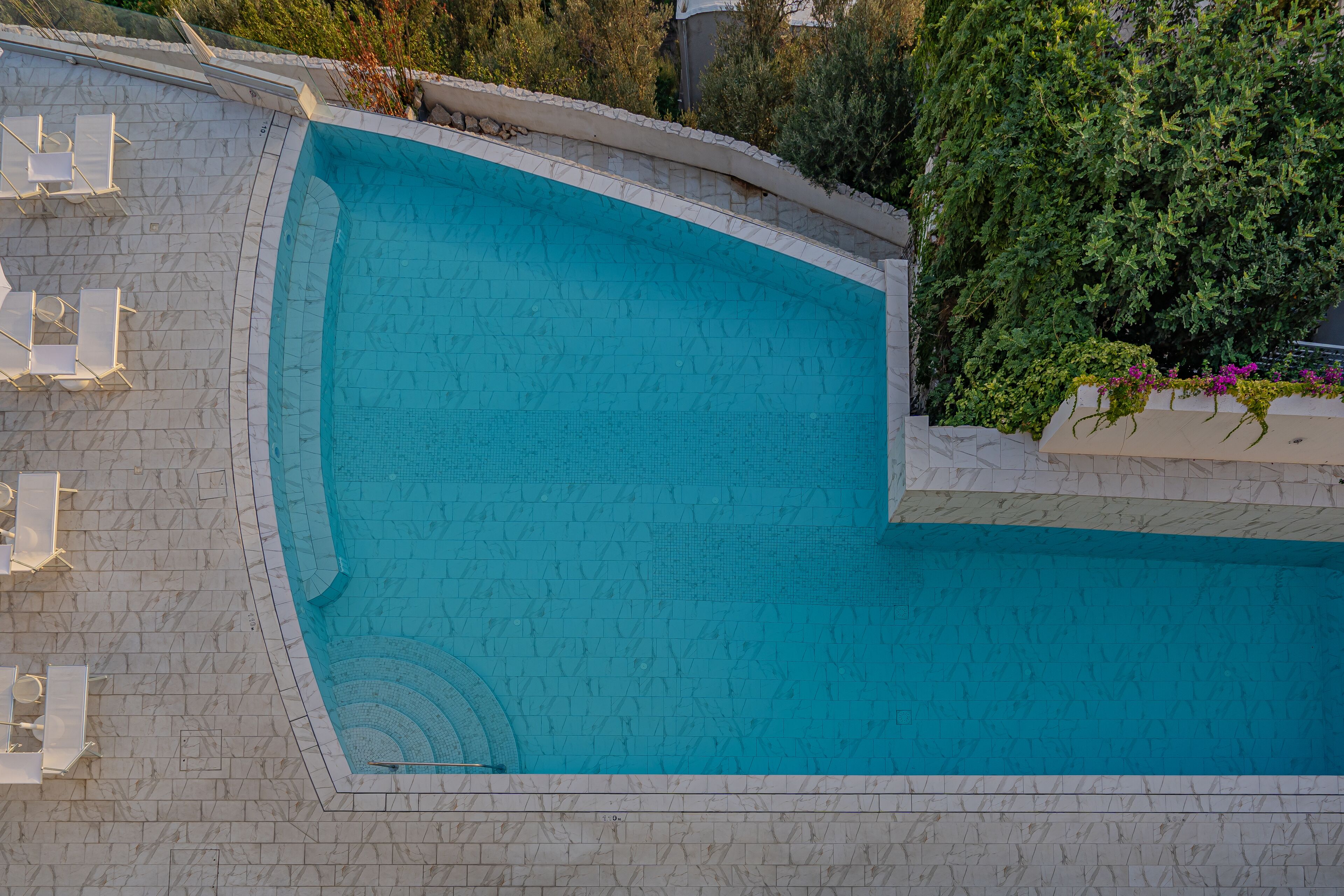 Une piscine à bord incurvé, entourée d’une terrasse carrelée et de chaises longues blanches.