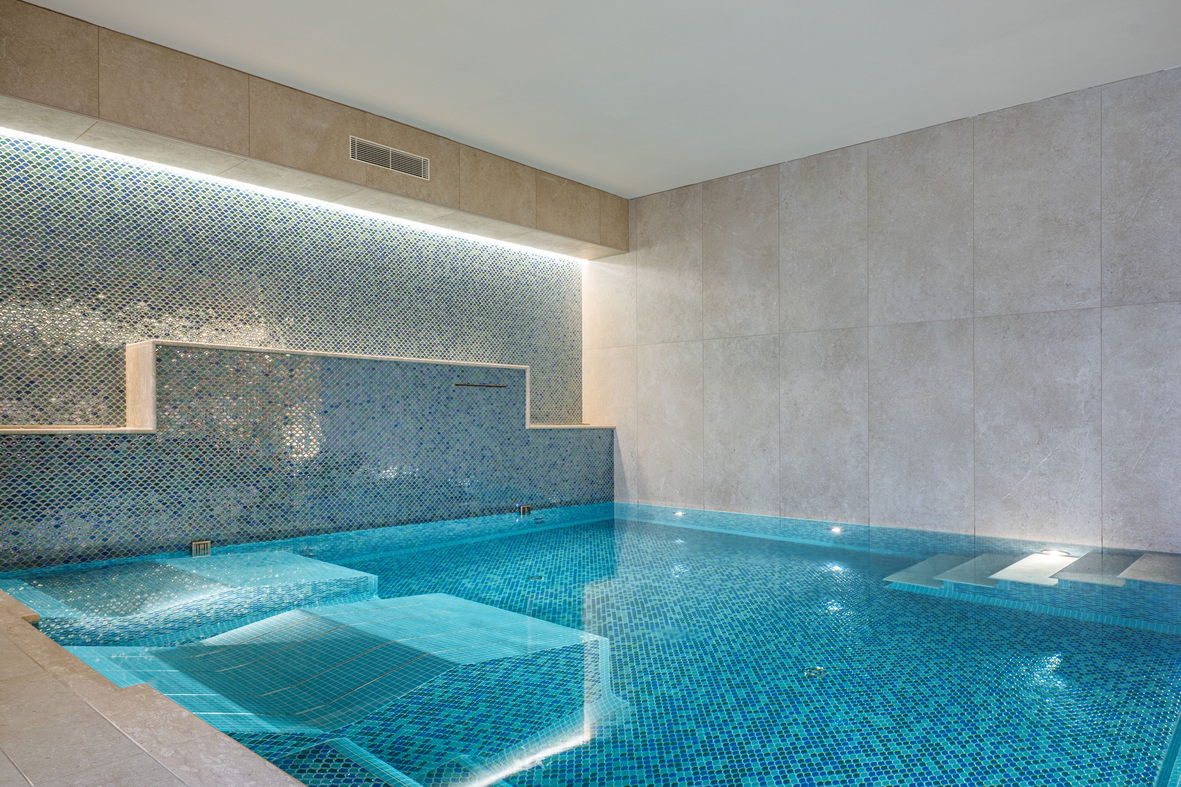 Ein moderner Innenpool mit klaren, blauen Mosaikfliesen, einer eingelassenen Sitzbank und einer wandbündigen Beleuchtung.