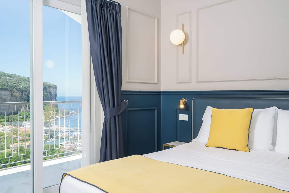 Una habitación de hotel con un ventanal grande, una cama con ropa de cama blanca y amarilla, una mesita de noche y una lámpara fijada a la pared.