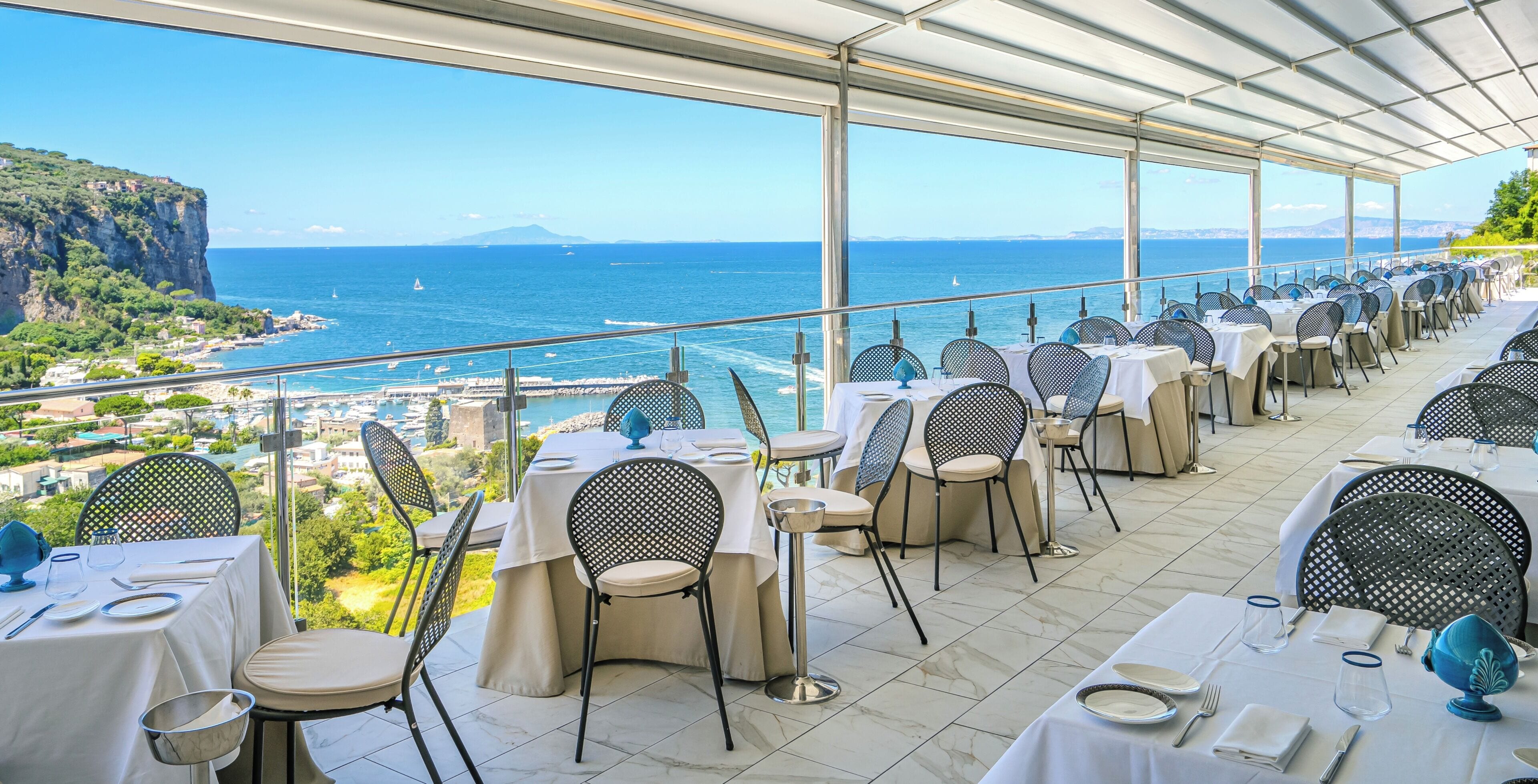 Terraço com mesas e cadeiras, com vista para a paisagem costeira com falésias e o mar.