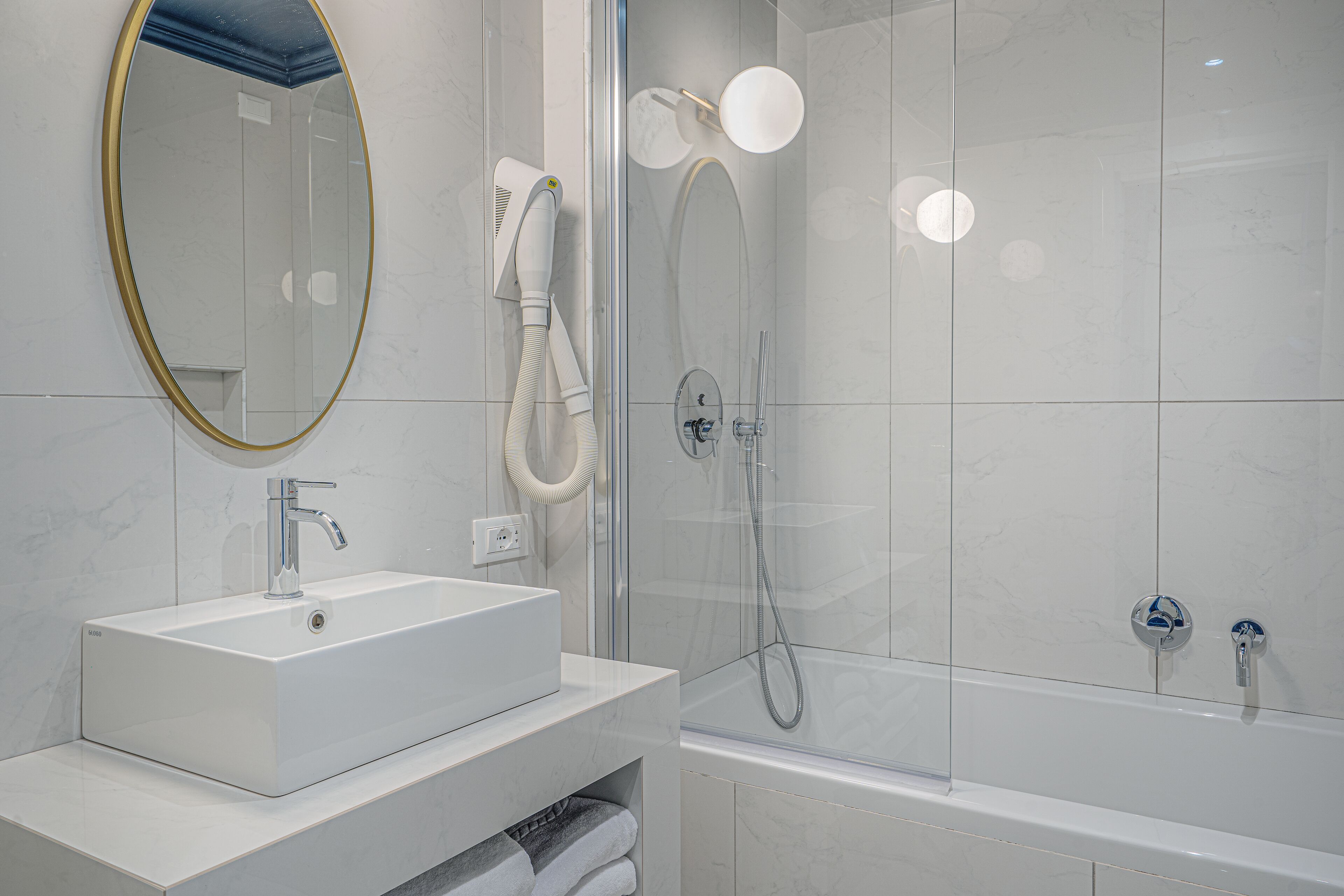 Un baño moderno con un lavamanos rectangular, un espejo con marco dorado, un secador de pelo portátil, una ducha con mampara de cristal y un cabezal de ducha manual, y una bañera.