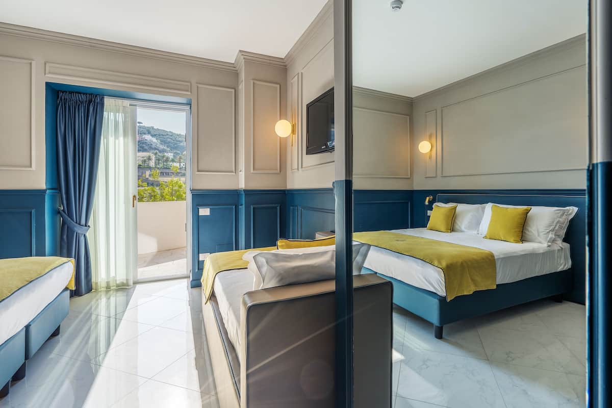 Habitación de hotel con una cama grande, una cama más pequeña y un sofá. Hay un televisor montado en la pared y una ventana con cortinas.