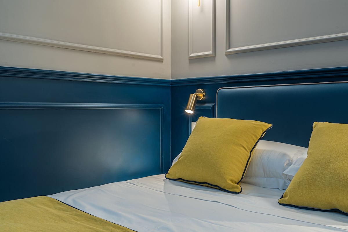 Una cama bien hecha con sábanas blancas y dos almohadas amarillo mostaza, una lámpara de noche y un cabecero azul.
