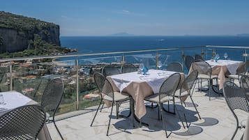 Un área de comedor al aire libre con mesas y sillas, con vistas a la costa, acantilados y el mar.