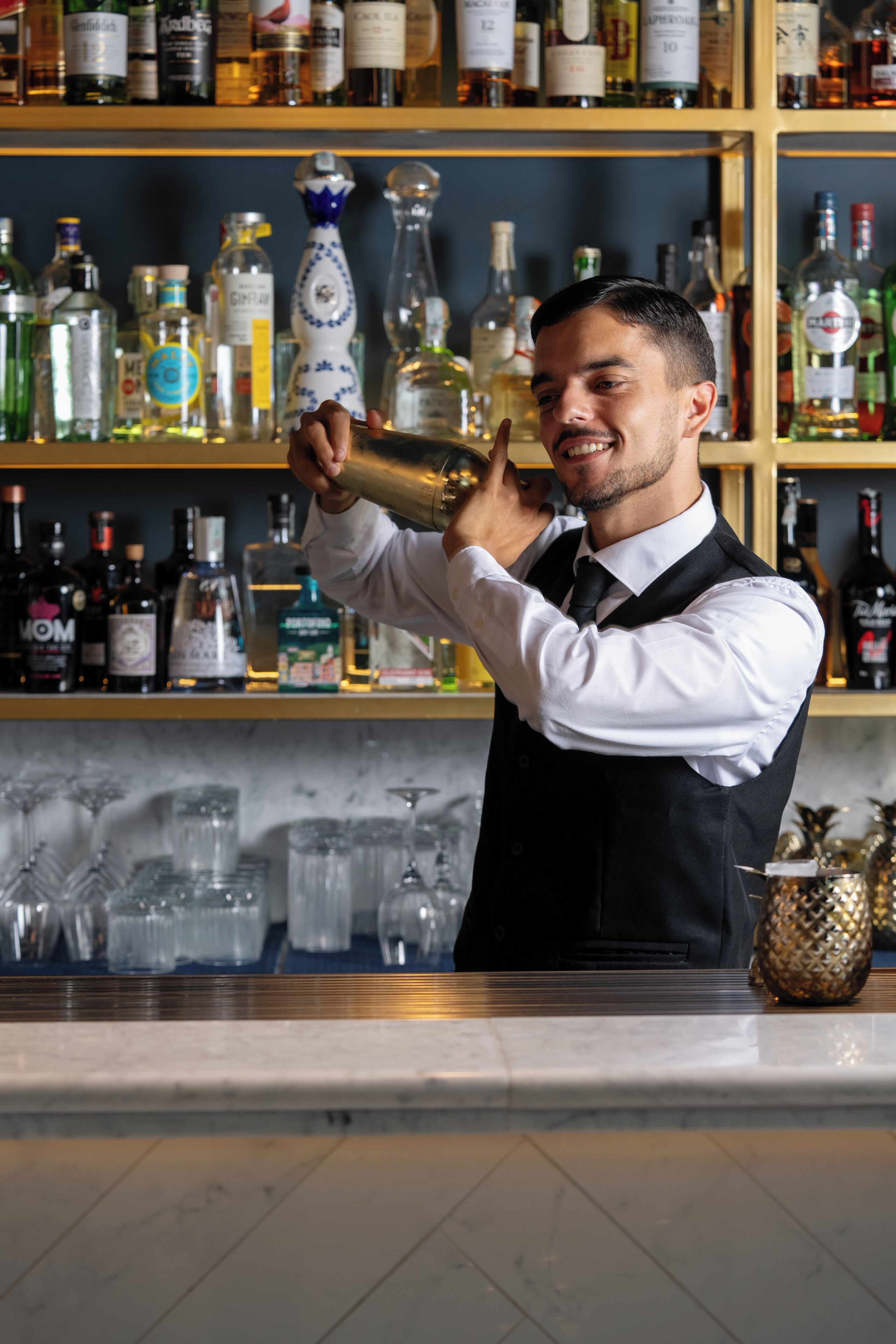 Um barman servindo uma bebida atrás do balcão, com prateleiras cheias de garrafas de bebidas.