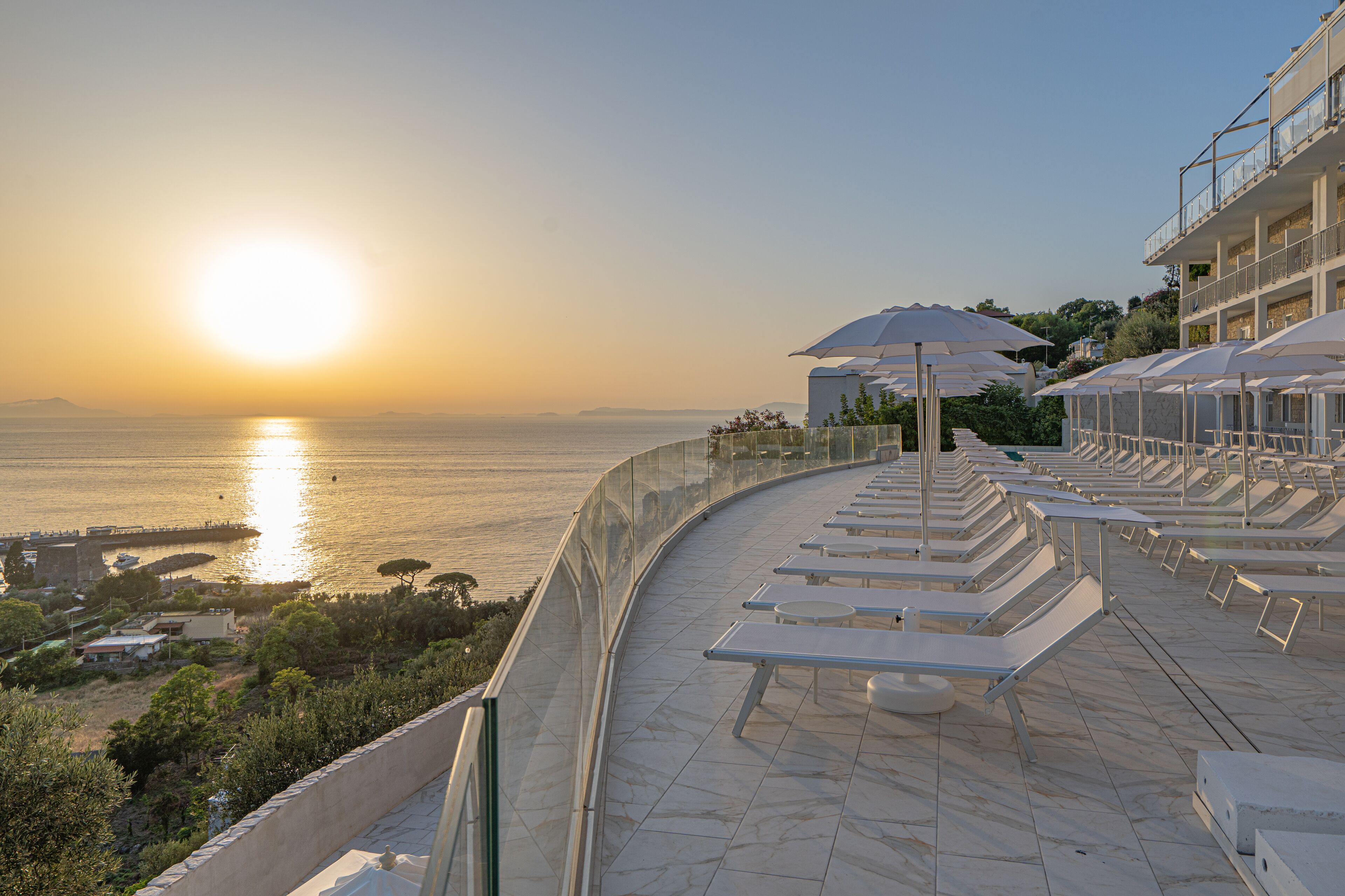 Una terrazza con lettini e ombrelloni affacciata su uno specchio d'acqua al tramonto.