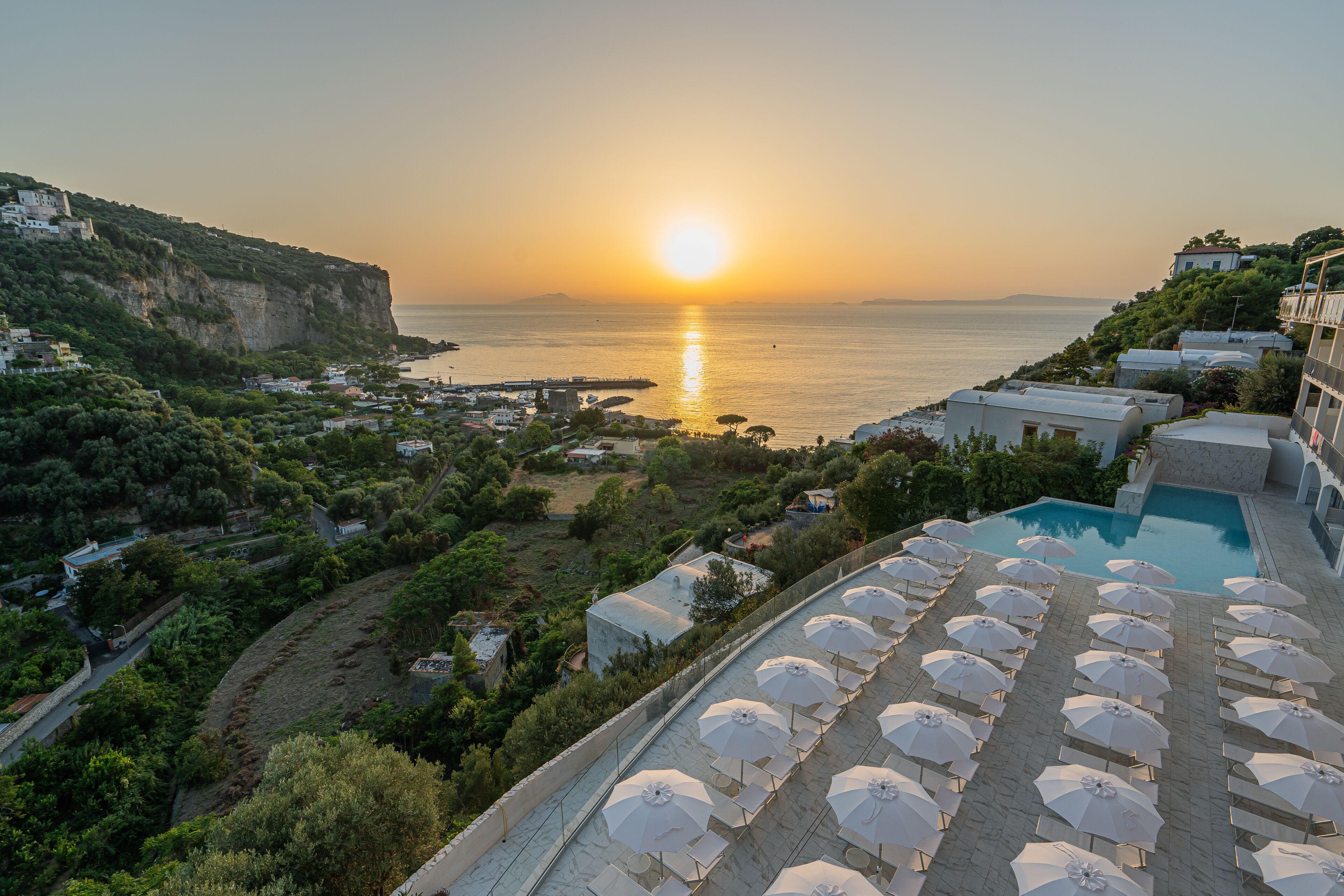 A resort with a pool area, sun loungers, and a view of the sea at sunset.