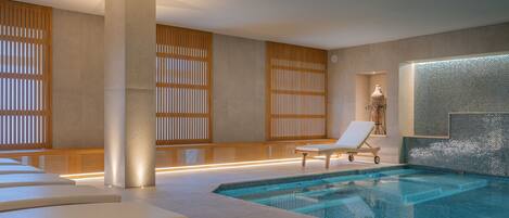 A modern indoor swimming pool with lounge chairs and a stone wall.