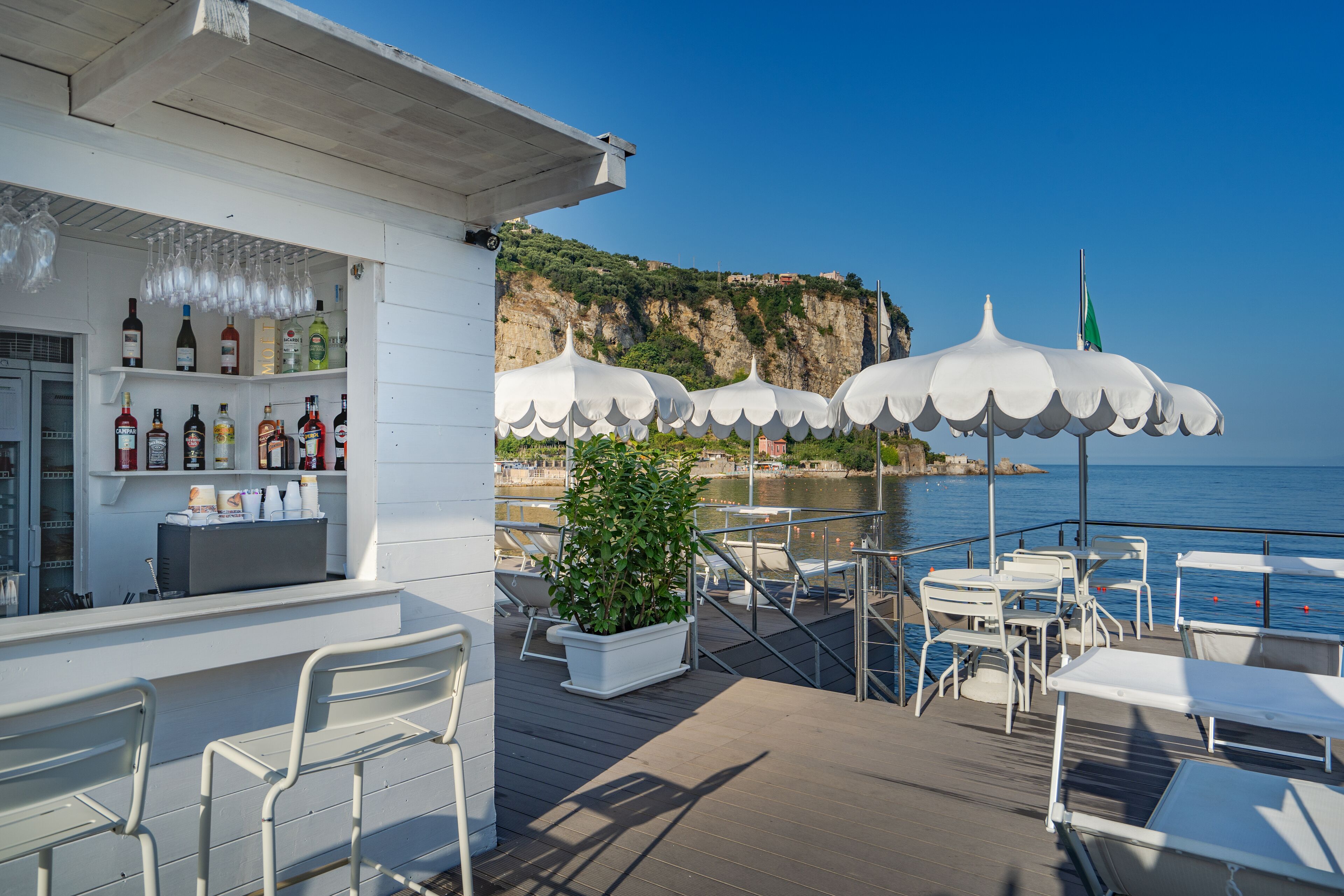 A deck with outdoor seating, a bar area with bottles, and a view of the sea and cliffs.