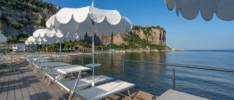 A deck with white lounge chairs and umbrellas, overlooking a calm sea and a rocky coastline.