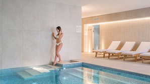 A swimming pool with tiled edges, a woman in a bikini standing by the pool, and lounge chairs in the background.