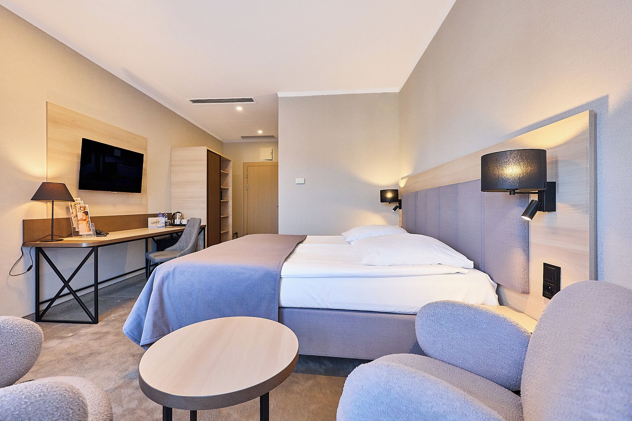 A hotel room with a bed, a desk, a chair, a round table, and a TV mounted on the wall.