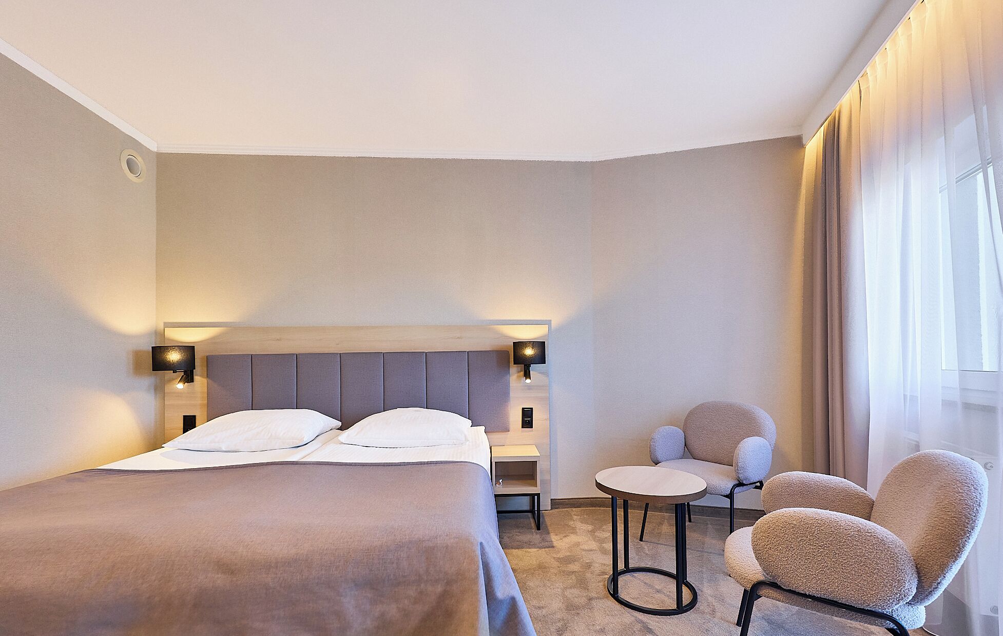 A hotel room with a bed, two chairs, a small table, and a window with curtains.