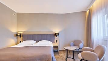 A hotel room with a bed, two chairs, a small table, and a window with curtains.