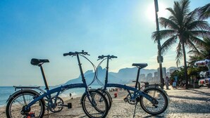Uma bicicleta estacionada numa praia de calçamento, com um horizonte urbano e palmeiras ao fundo.