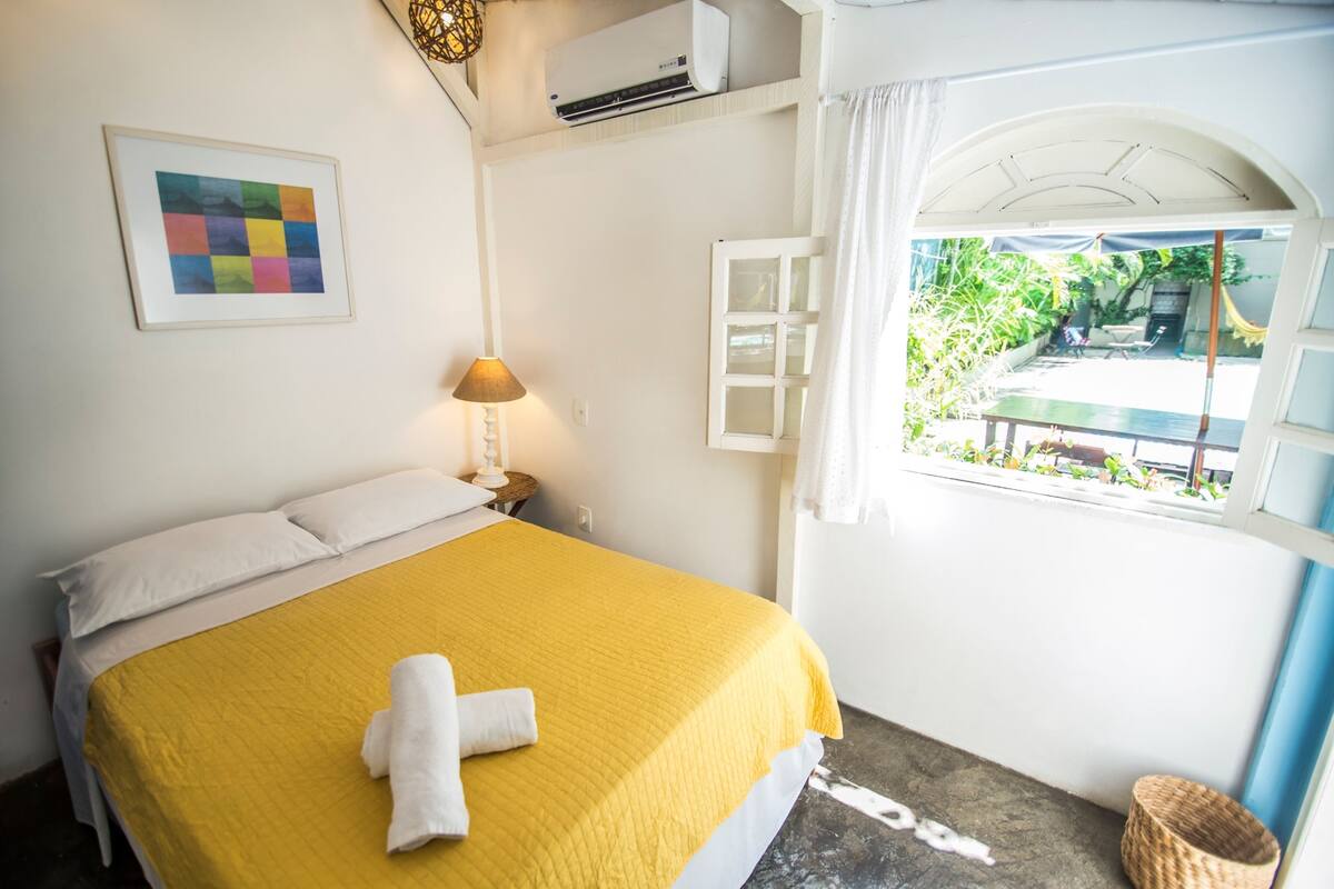 Quarto com cama, luminária de cabeceira, quadro emoldurado, ar condicionado e janela com vista para área verde.