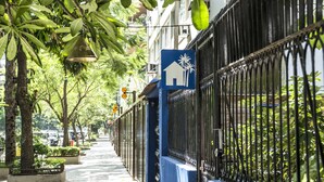Uma calçada arborizada com um prédio azul e um letreiro com o símbolo de uma casa em branco.