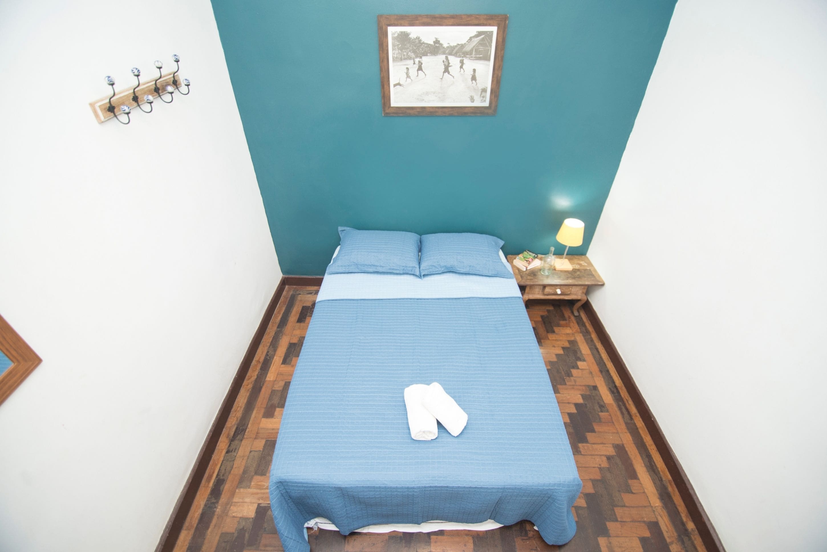 A neatly made bed with a blue bedspread and pillows, a wooden nightstand with a lamp, and a wall-mounted hookboard.