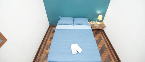 A neatly made bed with a blue bedspread and pillows, a wooden nightstand with a lamp, and a wall-mounted hookboard.
