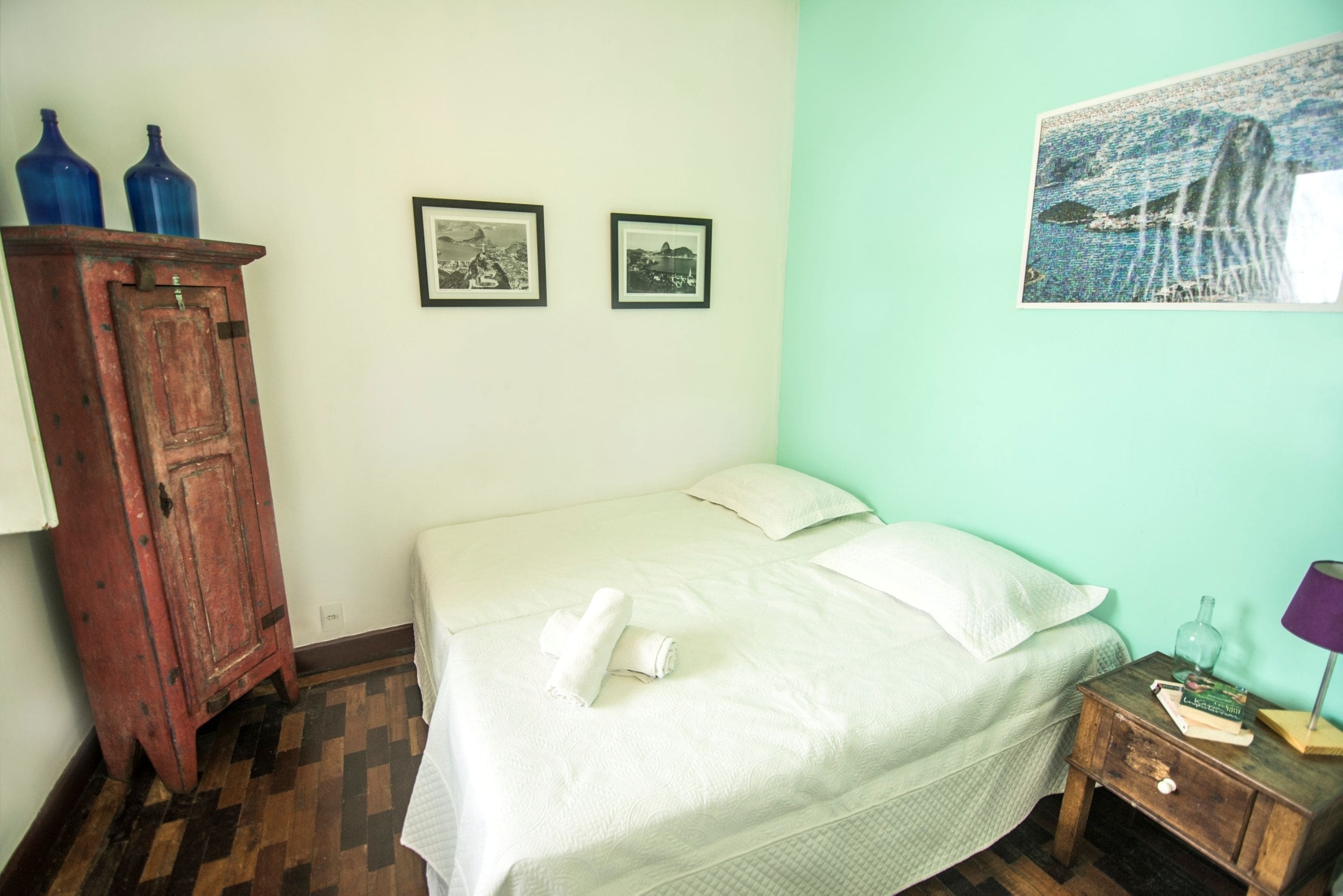 Quarto com cama, armário de madeira, duas molduras com fotos e uma mesa pequena com abajur.