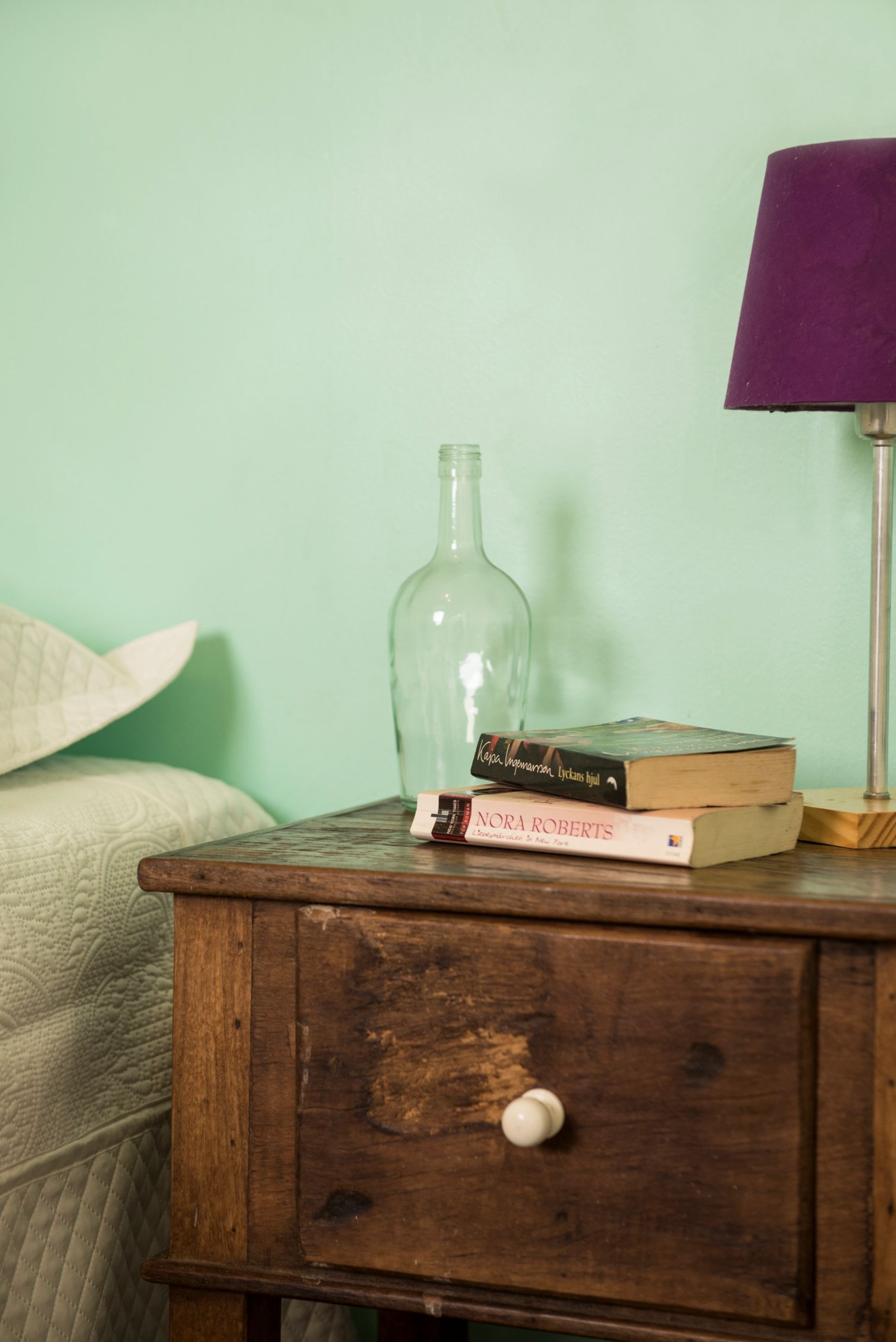 A wooden nightstand with a glass bottle, two books, and a lamp against a green wall.