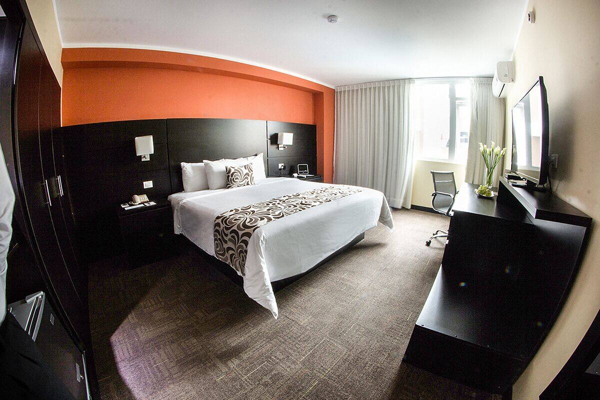 A hotel room with a large bed, a desk with a flat-screen TV, a wardrobe, a nightstand, and a window with curtains.