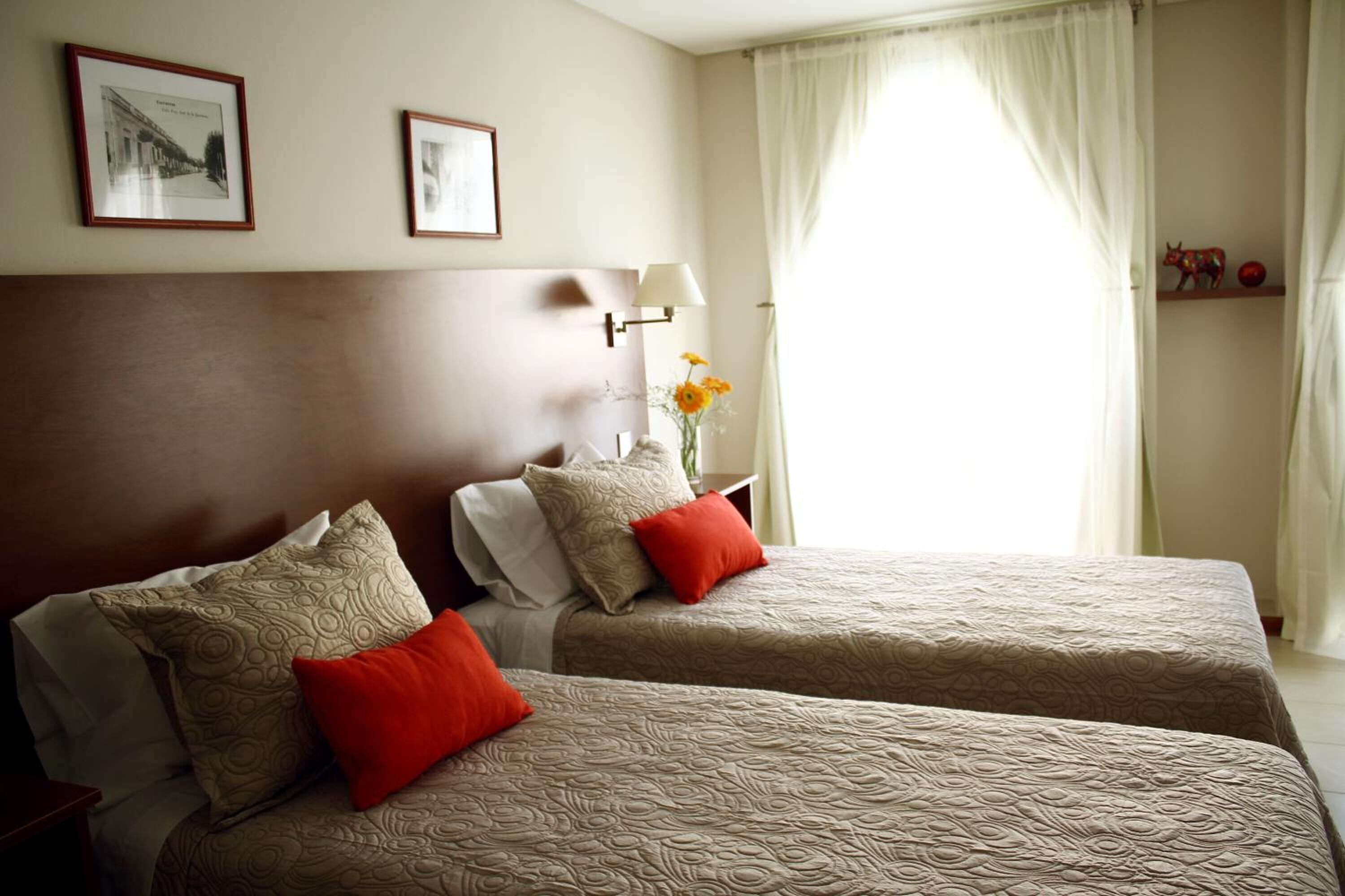 A hotel room with two beds, a nightstand with a lamp, and a window with curtains.