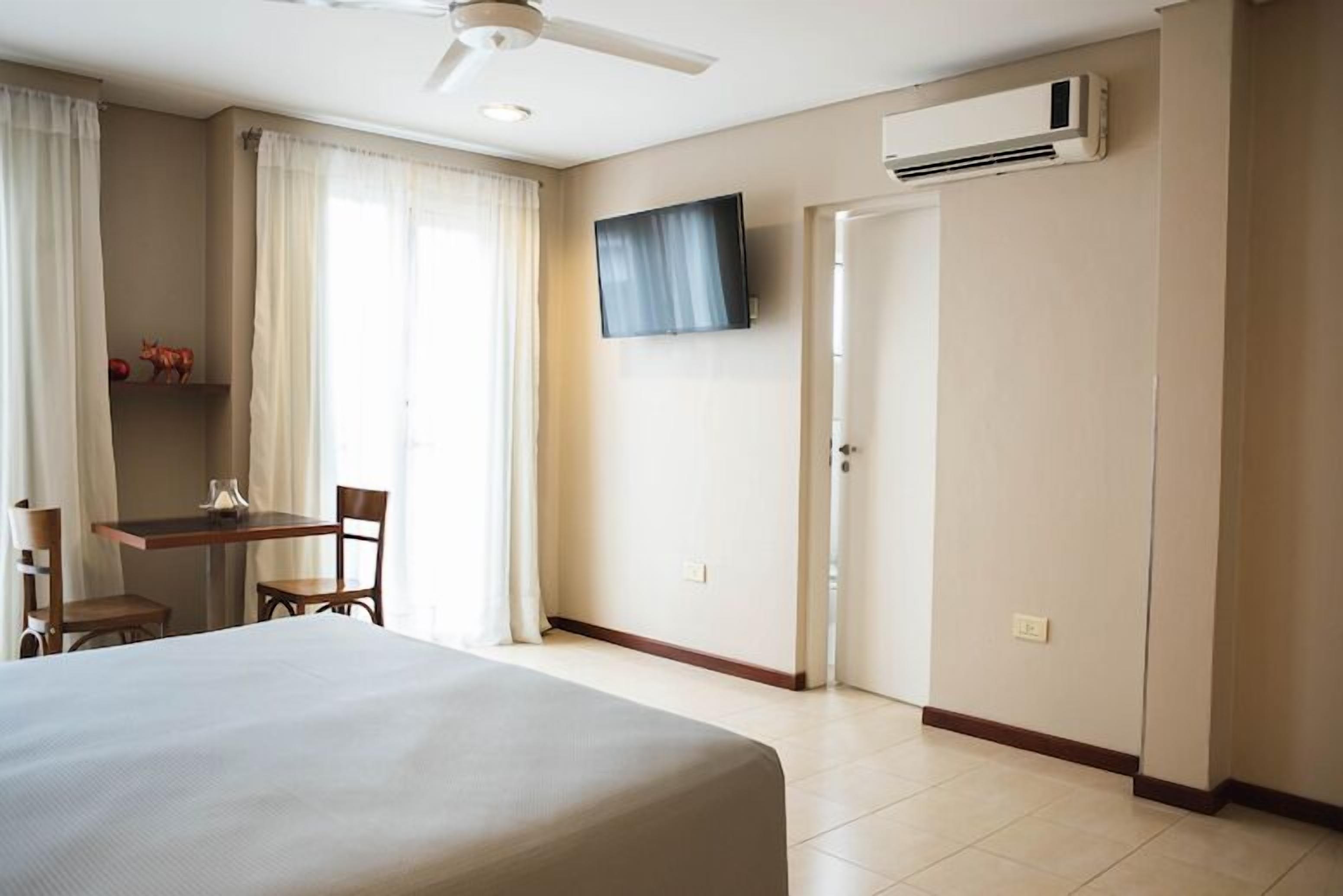 A hotel room with a bed, a desk with two chairs, a television, an air conditioning unit, and a door.
