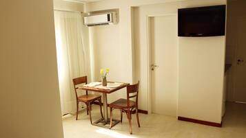 Quarto pequeno com uma mesa de jantar, duas cadeiras, um vaso com flores, aparelho de ar condicionado, uma porta e uma televisão na parede.