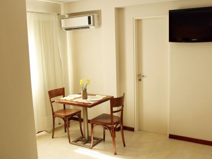 Quarto pequeno com uma mesa de jantar, duas cadeiras, um vaso com flores, aparelho de ar condicionado, uma porta e uma televisão na parede.