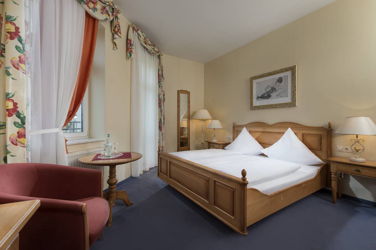 A hotel room with a wooden bed, two bedside lamps, a small round table with a bottle, a chair, and a window with curtains.