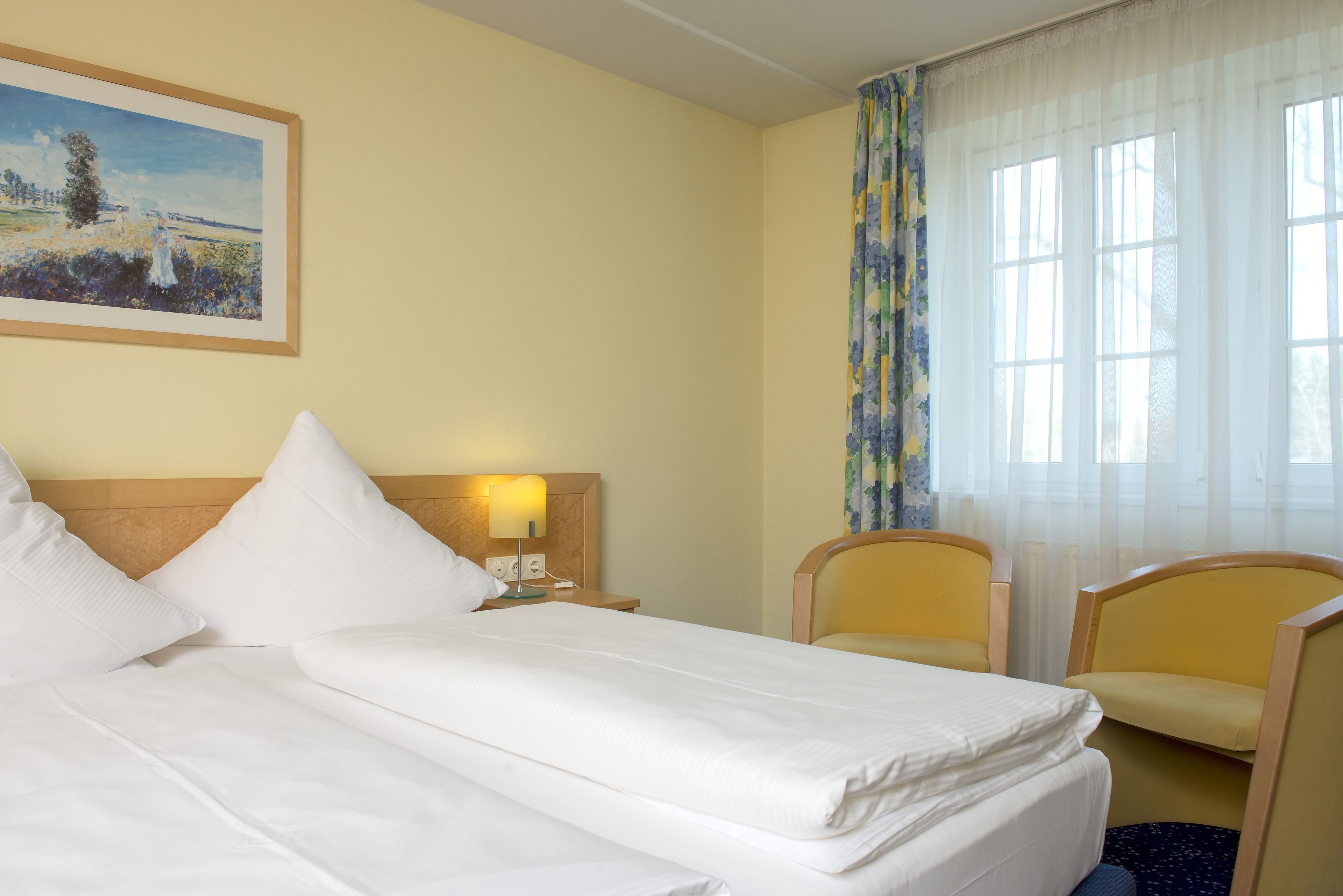 Habitación de hotel con cama, dos sillas, una mesita de noche con lámpara y un cuadro en la pared.