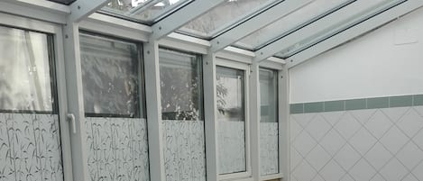 A sunlit outdoor seating area with white lounge chairs and a table, enclosed by a glass roof with patterned windows.