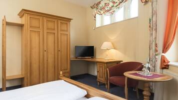 Ein Hotelzimmer mit zwei Betten, einem Holzschrank, einem Schreibtisch mit Fernseher, einem Sessel, einem kleinen Tisch mit Lampe und einem Fenster mit Vorhängen.