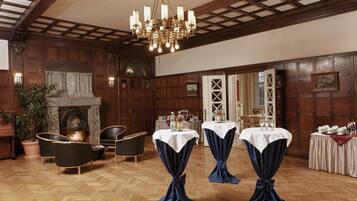 Ein Zimmer mit Kamin, Holzwänden und einer Chandelüster. Es gibt Tische mit weißen Tischdecken und blaue Vorhänge.