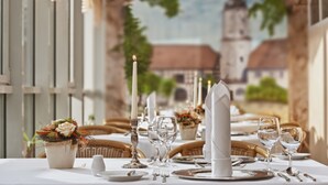 Mesa para comer con mantel blanco, cubiertos y vasos transparentes, una vela y un centro de mesa floral.