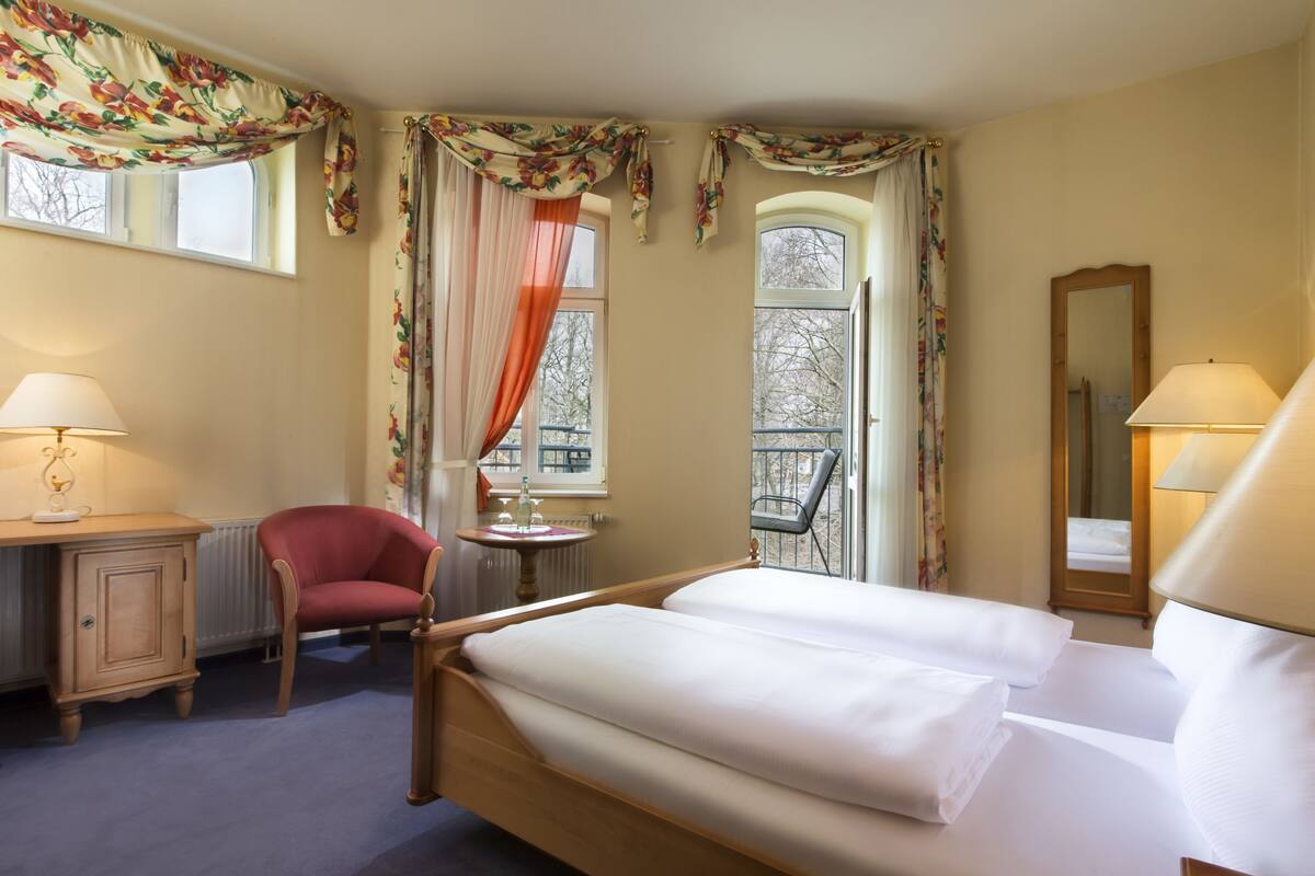 A hotel room with a bed, a desk, a chair, a small table, and a window with curtains.