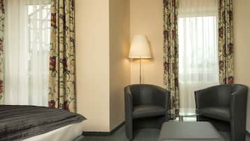 A hotel room with a bed, two armchairs, a small glass table, a floor lamp, and floral curtains.