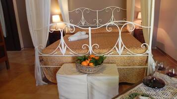 Quarto bem arrumado com cama, mesas de cabeceira, uma mesinha com frutas e copos de vinho, e um prato decorativo.