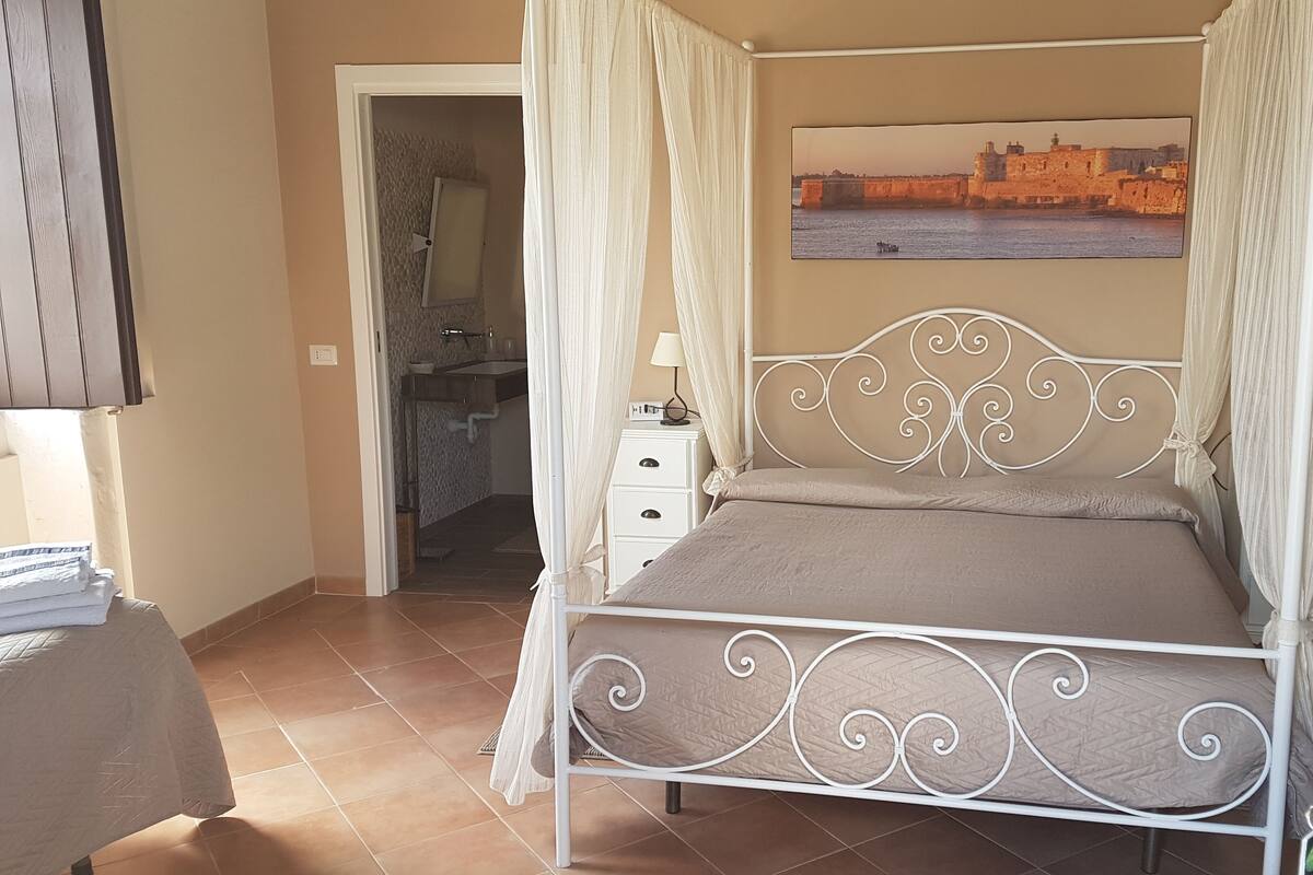 Un dormitorio con cama, mesita de noche, televisor montado en la pared y un baño visible a través de una puerta abierta.
