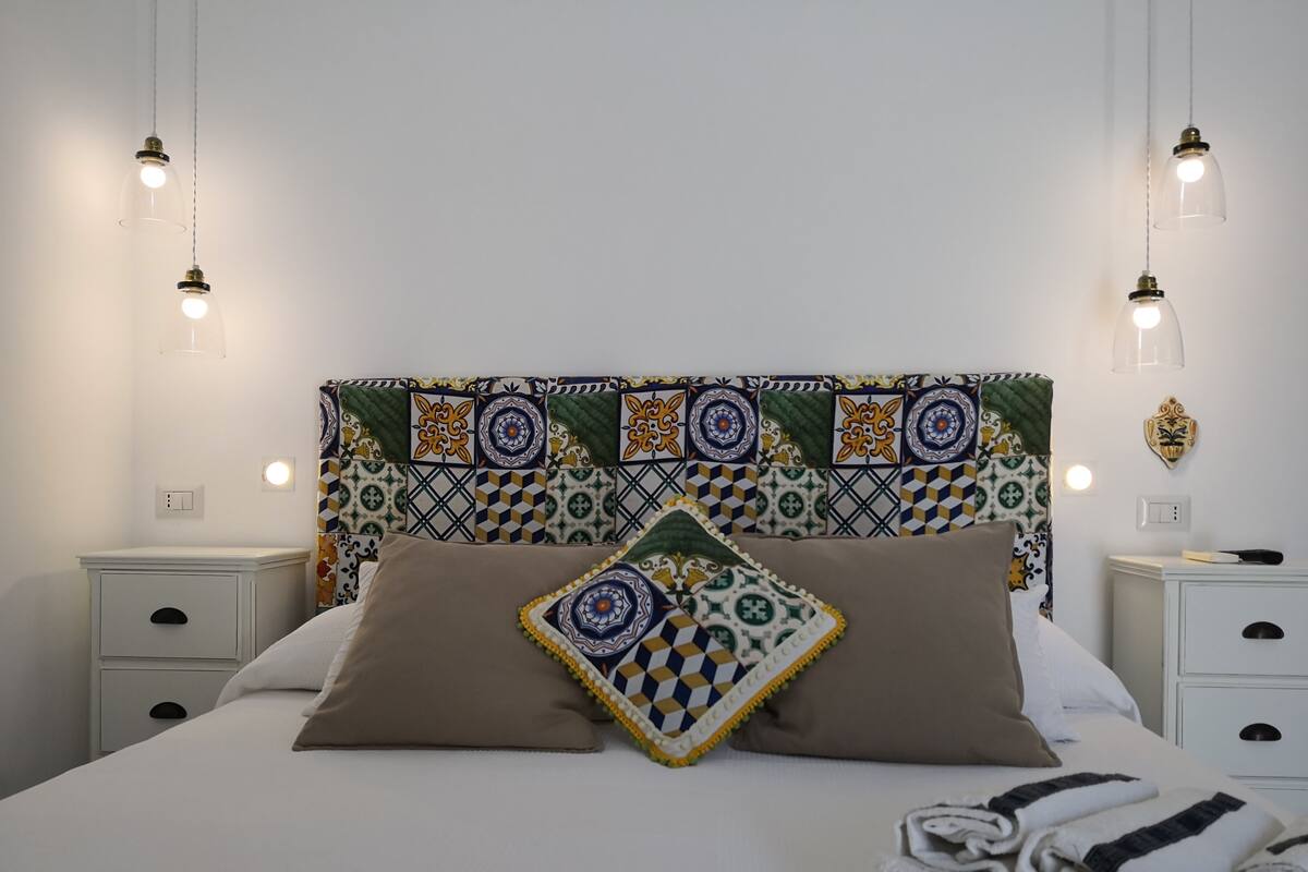Una cama bien tendida con un cabecero estampado, mesitas de noche blancas y una pared con un diseño decorativo de azulejos.