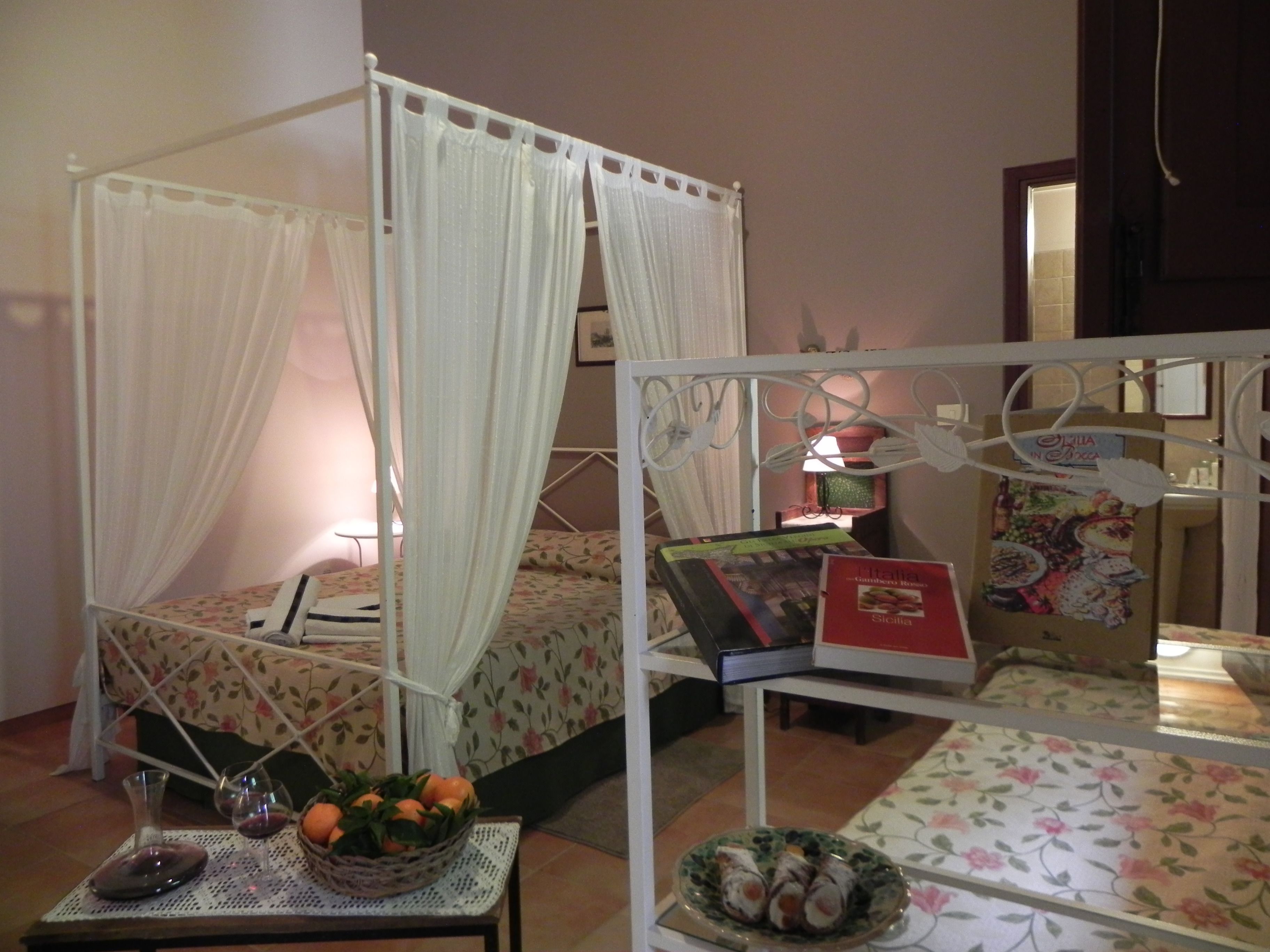 A bedroom with a canopy bed, a table with a basket of oranges and wine glasses, and a decorative plate with food items.