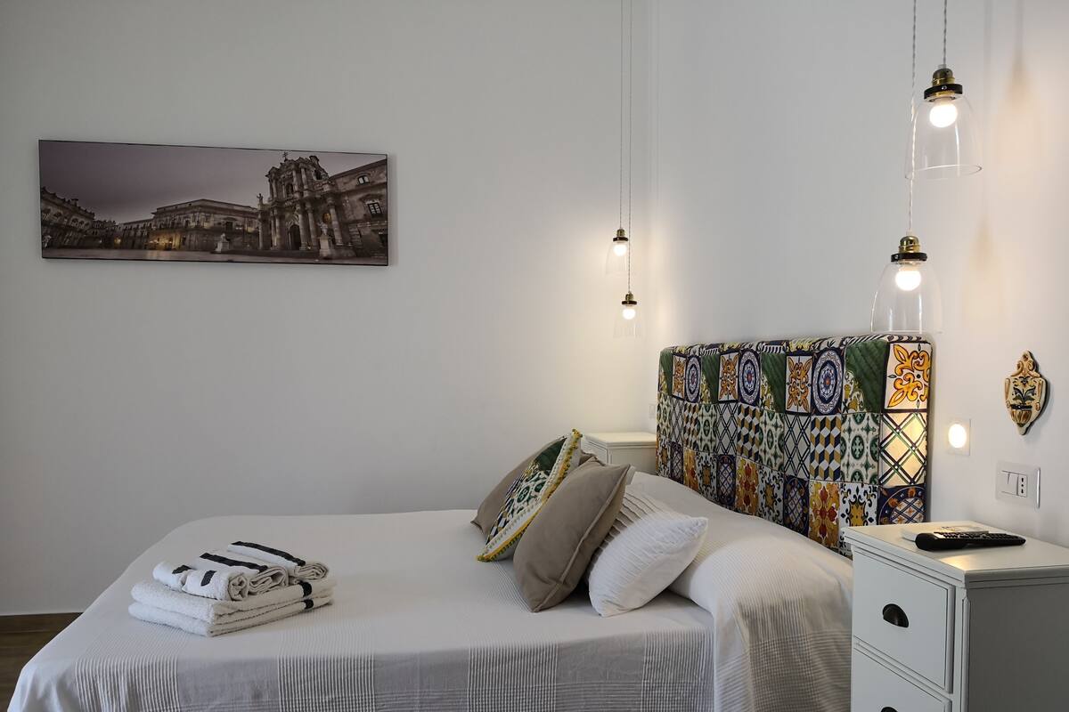 Una cama bien hecha con toallas dobladas, un cabecero estampado, una mesita de noche con un control remoto y una lámpara de pared.
