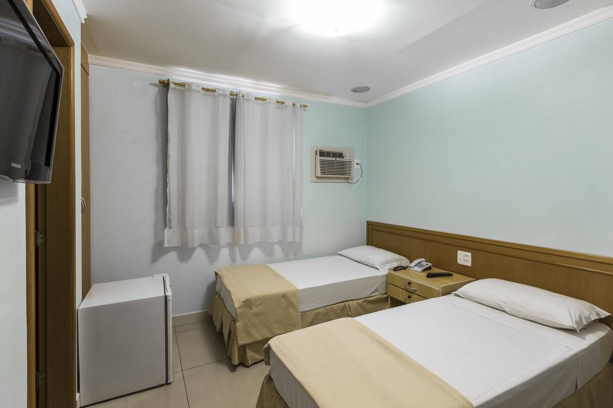 A hotel room with two beds, a small refrigerator, a bedside table with a phone, and a wall-mounted air conditioner.