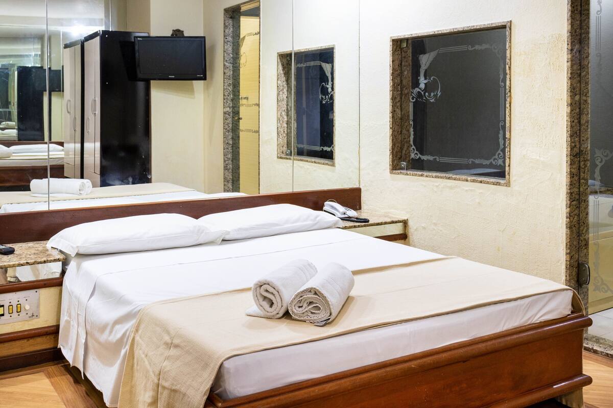 A hotel room with a bed, a bedside table, a mirror, a TV, and a window with decorative frames.
