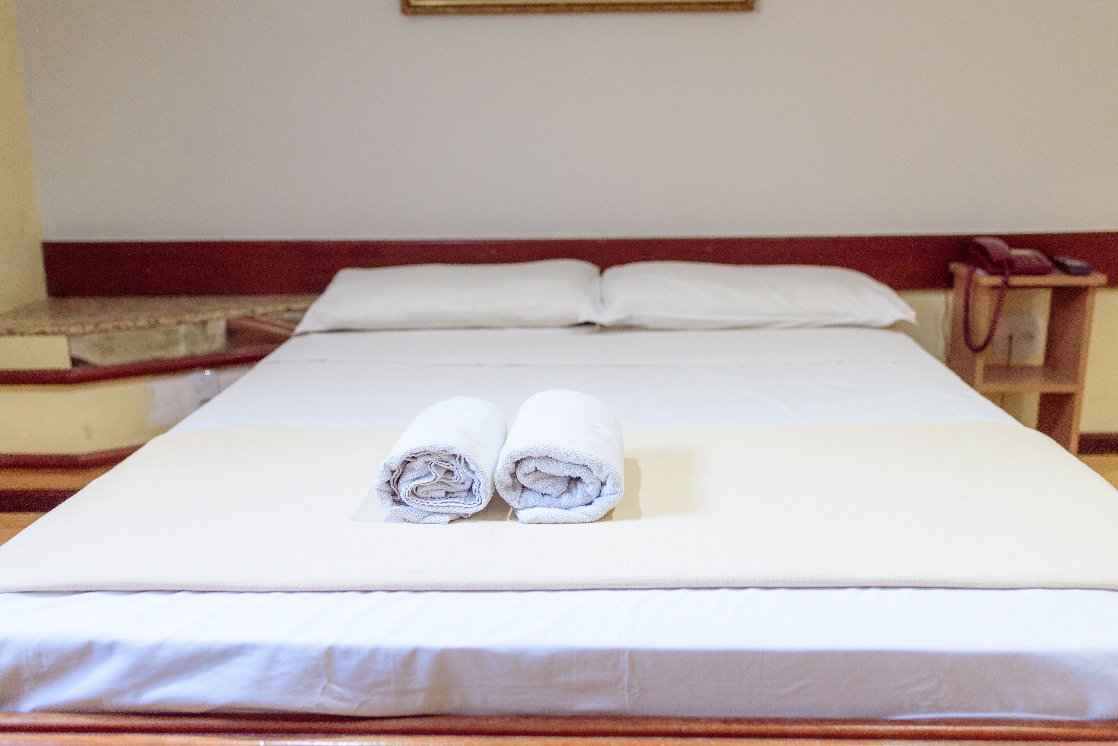 A neatly made bed with two rolled towels, a wooden headboard, and a bedside telephone.